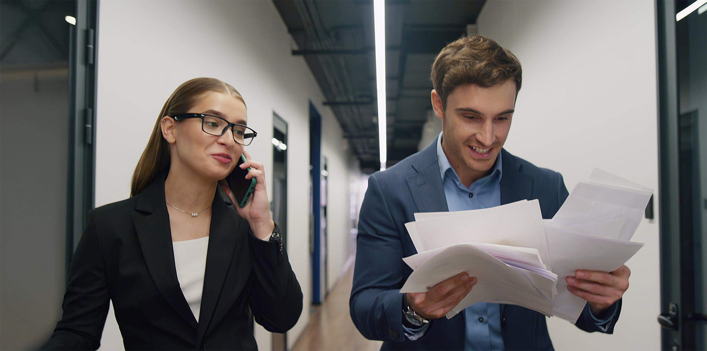 Mies ja naistyöntekijä toimiston käytävällä tutkimassa papereita, nainen puhuu puhelimessa