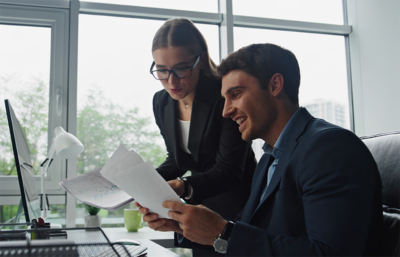Kaksi toimistotyöntekijää tutkimassa papereita tietokoneen ääressä